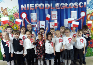 Dzieci z grupy 5-6-letniej stoją w strojach galowych na tle napisu "Dzień Niepodległości", trzymają w rękach flagi Polski.."